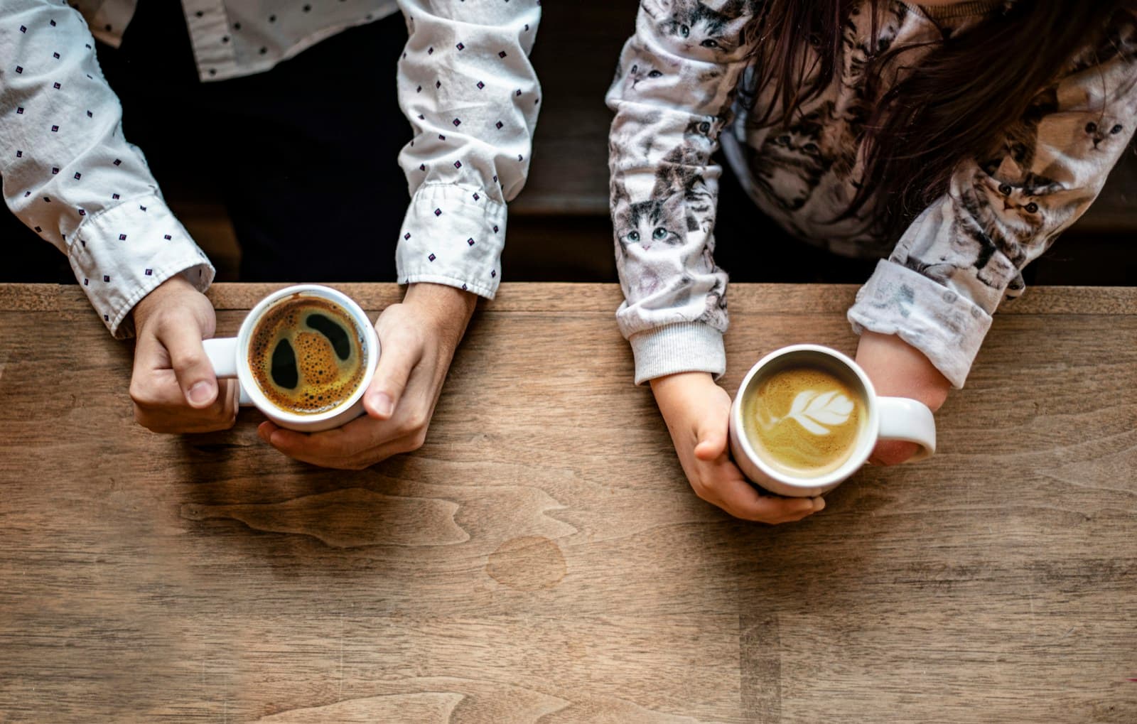 Two people's hands round cups of coffee on a wooden table